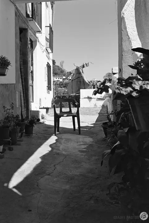 A black-and-white photo of a solitary chair in a narrow passage. A scarecrow is visible in the garden behind.