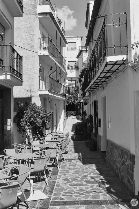 A black-and-white photo of a narrow street in Soportújar with empty cafe tables and chairs along one side (it's siesta time).