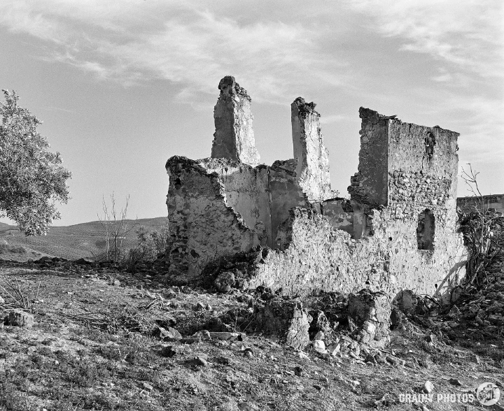 A black-and-white photo of the ruins of an abandoned farmhouse. Only some of the stone walls are standing. There is no roof or windows and doors.
