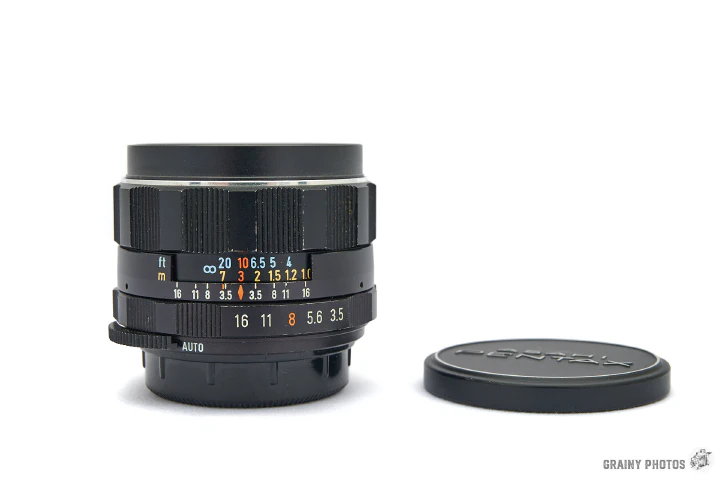 A photo of the Super-Takumar 28mm f3.5 lens stood vertically with a lens cap next to it.