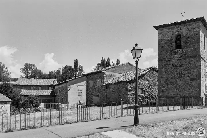 A black-and-white film photo of houses in Alba de los Cardaños with the Iglesia de San Justo y Pastor bell tower on the right