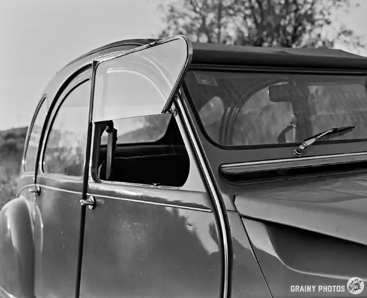 A black-and-white film photo of the side of the car, in particular the front door with an open window. The window hinges upwards on itself to open.