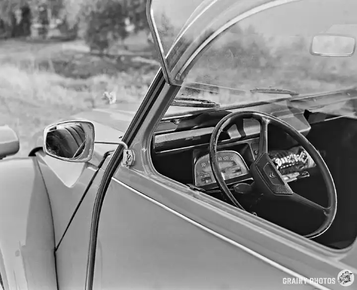 A black-and-white film photo looking in through the open driver's window. The instrumentation is very sparse - mostly just a speedometer is visible.