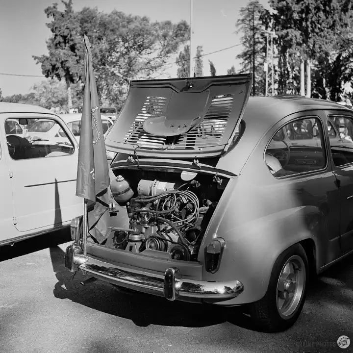 Black-and-white film photo of a with its rear engine bay cover open exposing the engine. There is a flag fixed to the rear bumper.