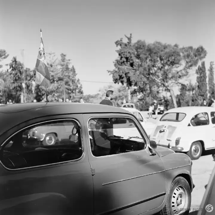 Black-and-white film photo of several Seat 600 cars at the show. The nearest has a flag fixed to the roof.