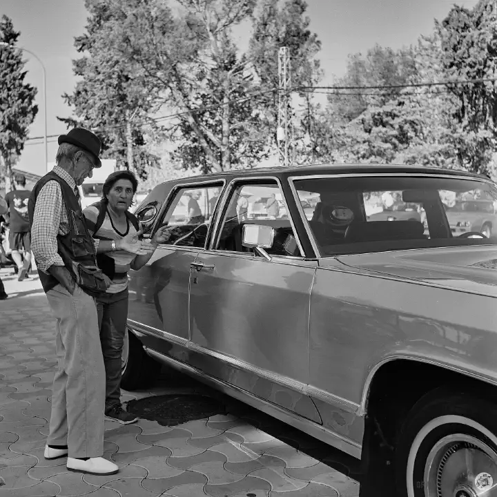 Black-and-white film photo of a man and woman next to an American car. The lady has a startled expression.