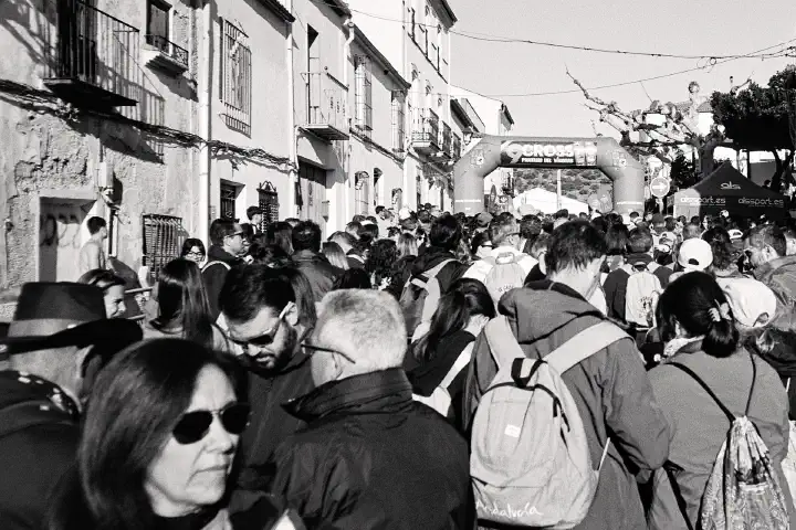 A crowded street scene features a mix of people gathered hikers gathering for the start of the walk, many wearing coats and backpacks. A large inflatable archway with text marks the finish line of the annual race. The street is lined with old buildings. Black and white image.