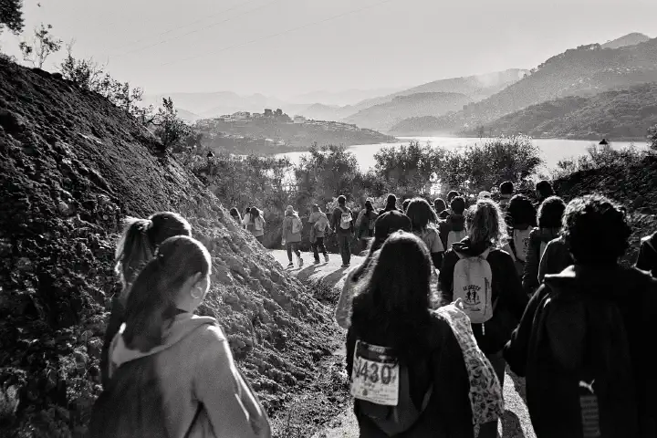 A group of people wearing backpacks and numbered bibs walk along a dirt path near Las Casillas de Martos. The landscape features rolling hills, a distant village, and the shimmering Viboras Reservoir under a clear sky.