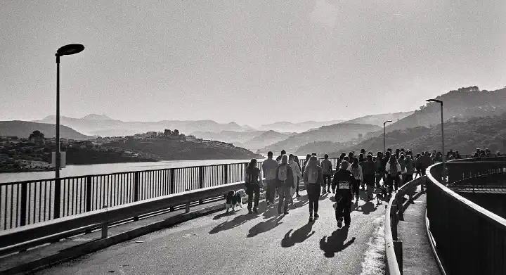 A group of people and a few dogs walking across the dam during the Cross Pantano del Víboras. The black-and-white scene captures the hills and water of the Viboras Reservoir in the background, their outlines softened by a slightly hazy sky.