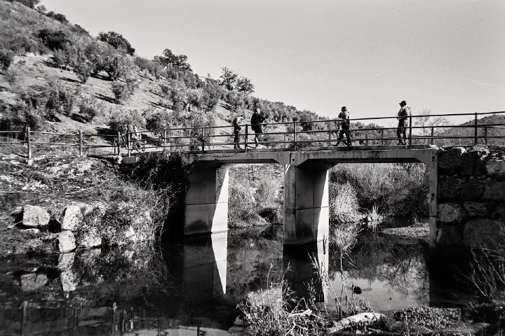 Black and white photo of a rural scene on the Pantano del Víboras course, with three people walking across a small bridge over a calm stream. The bridge has railings, and rocky hills with sparse trees reflect in the water below.