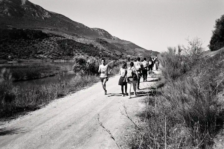 One of the leading runners passes the walkers going in the opposite direction on a dirt track by the reservoir. The landscape features hills and sparse vegetation under a clear sky. The image is in black and white.