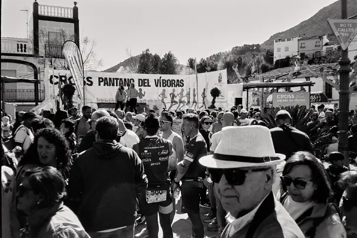 A large crowd gathers in the Las Casillas de Martos village square after the Cross Pantano del Víboras event. People, some in hats and running gear, mill about. In the background, there is a stage for the presentation ceremony.