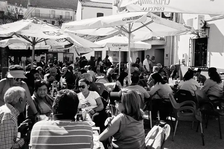 A bustling outdoor street party scene in black and white, with many people seated under umbrellas, chatting and enjoying food and drinks. The atmosphere is lively and social, with a backdrop of white buildings and sunlight filtering through the umbrellas.