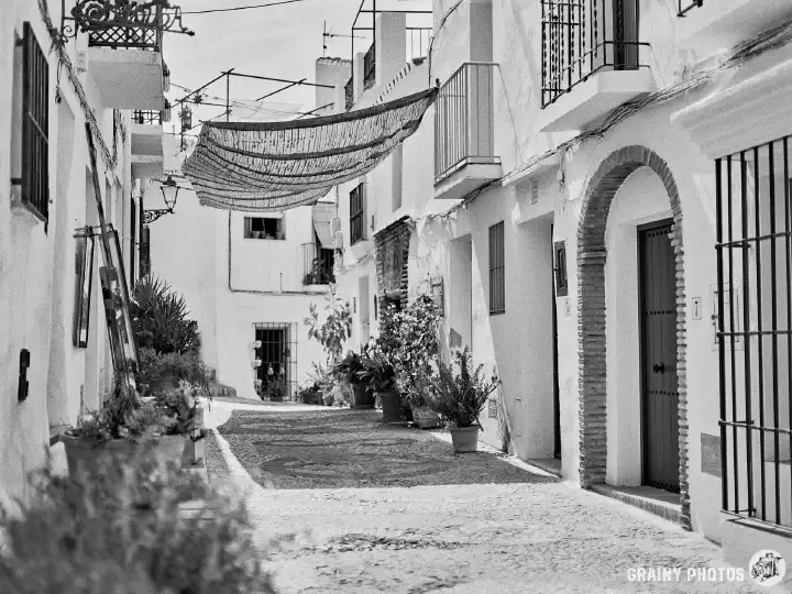 A black-and-white photo of a pretty pebble-patterned narrow street with pretty white terraced houses. A sun screen is stretched across the street for shade.