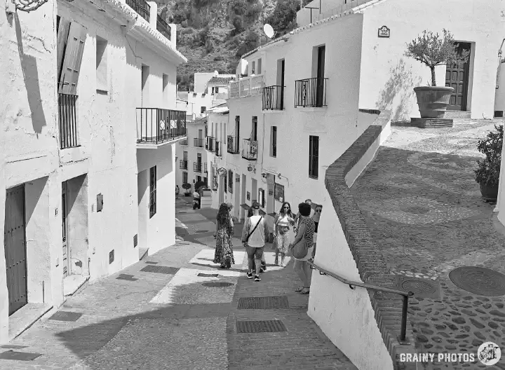 A black-and-white photo of a pebbled split-level narrow street with terraced white houses. A small group of tourists are in the lower street.