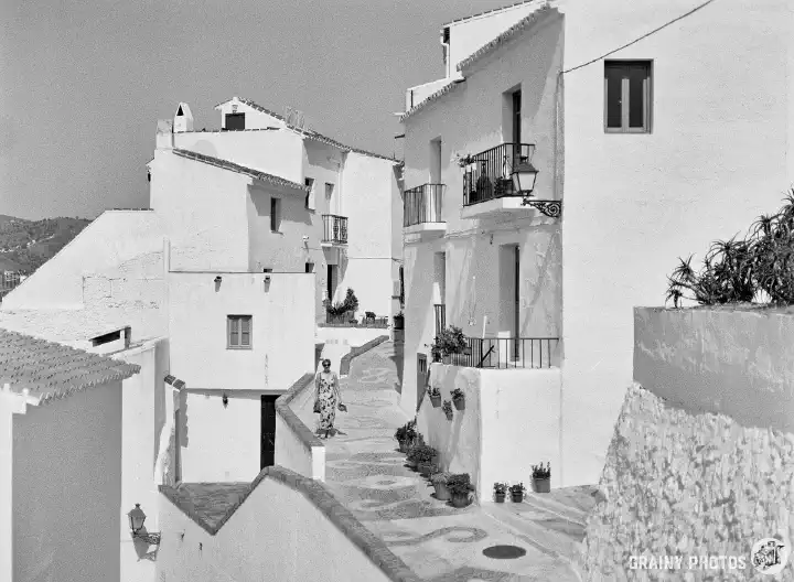 A black-and-white photo of a solitary tourist wandering along the patterned pebble narrow streets amongst the white houses.