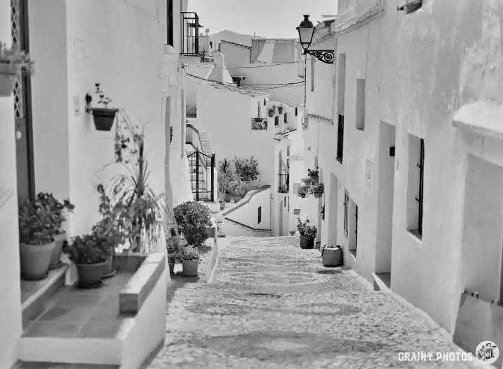 A black-and-white photo of a narrow pebbled street going steeply downhill, disappearing from view. White houses on both sides. Numerous potted plants are in the street in front of the houses.