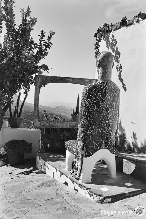 A black-and-white photo of a sculpture of a very tall man sitting on a stool watching TV. The TV is just a frame, with a scenic landscape visible through it.