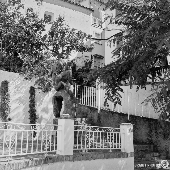 A black-and-white photo of a wooden sculpture on a small terrace beside some steps. White houses behind and to the right with several trees. A very picturesque setting.