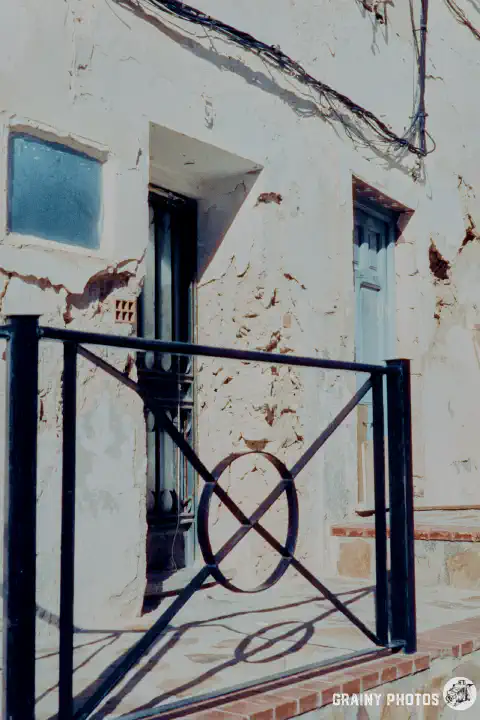 A colour film photo of the front doors to two terraced white houses. The render has spalled extensively, and the paint is peeling. Shot on Harman Phoenix 200 film.
