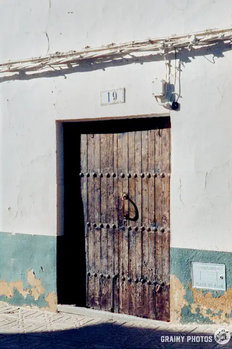 A colour film photo shot on Harman Phoenix 200 film. An old timber front door. The house number (19) is above the door. The lower part of the wall is painted green, with peeling paint. The upper part of the wall is white.