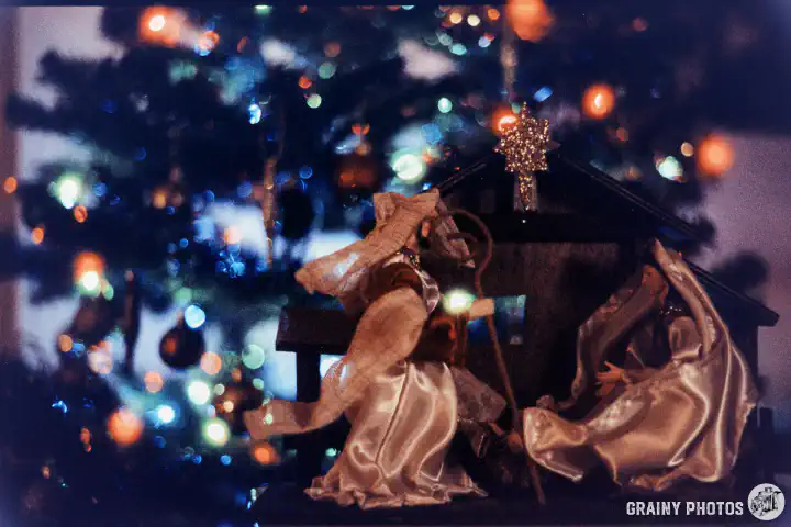 A colour film photo of a belén in front of a Christmas tree. The Christmas tree is out of focus and the coloured lights appear as coloured bokeh. Shot on Harman Phoenix 200 film.