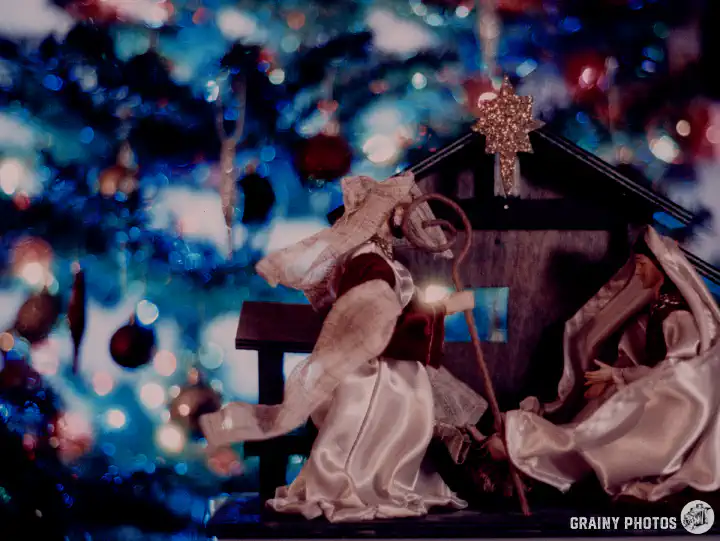 A colour film photo of a belén in front of a Christmas tree. The Christmas tree is out of focus and the coloured lights appear as coloured bokeh. Shot on Harman Phoenix 200 film.