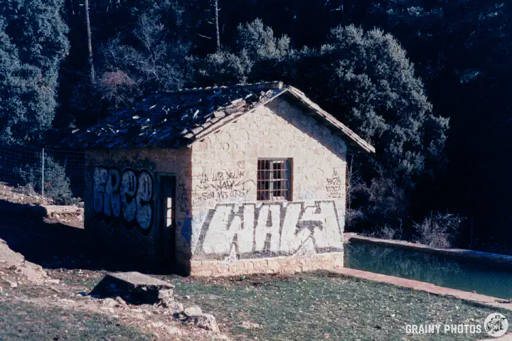 A colour film photo of an abandoned stone hut by a pool. Graffiti has been sprayed on the two visible walls.