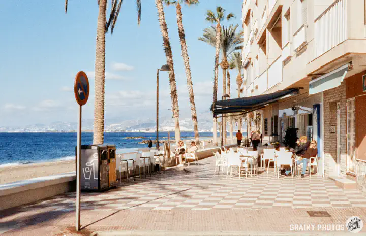 A colour film photo of cafes on a paved promenade with a terrific sea view.