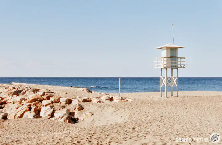 A colour film photo looking out to sea. In the foreground is a deserted beach with a rocky outcrop and a lifeguard lookout hut on the right.