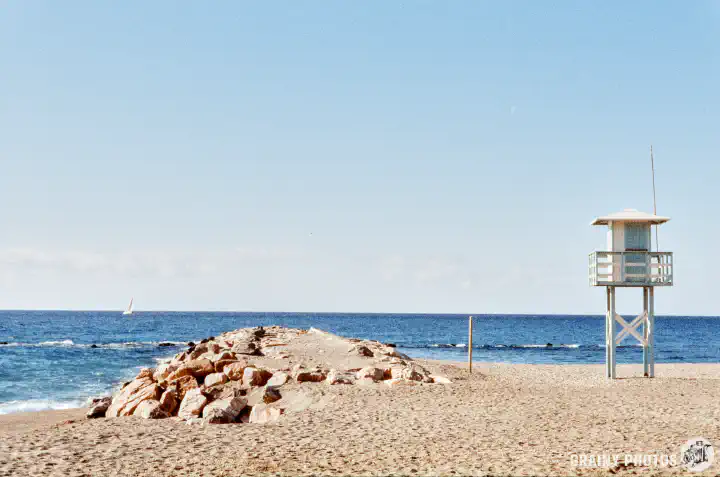 A colour film photo looking out to sea. In the foreground is a deserted beach with a rocky outcrop and a lifeguard lookout hut on the right. On the sea horizon, there is a sailing boat.
