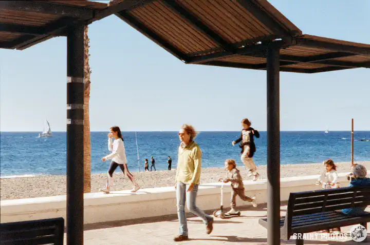 A colour film photo of people walking along the seafront on the promenade. The main subjects are framed by posts supporting a sunshade. Two sailing boats are visible out at sea in the distance.
