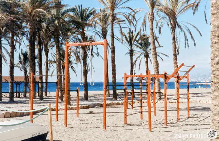 A colour film photo of a children's play area with swings and climbing frames on the beach amongst palm trees.
