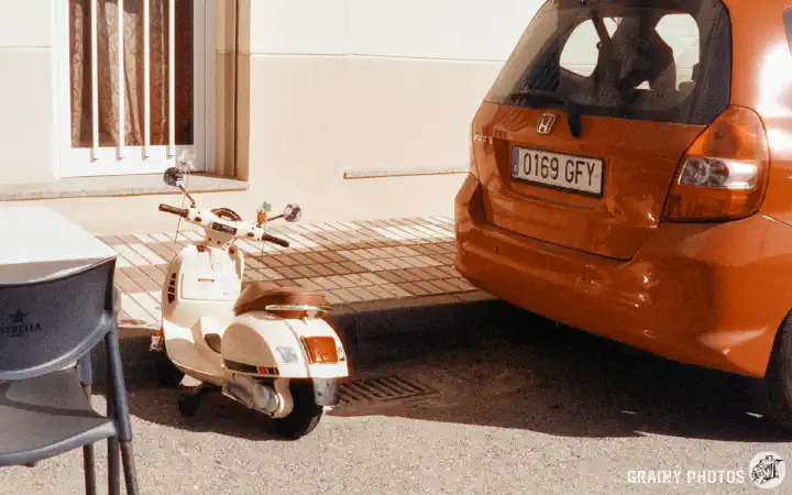 A colour film photo of a tiny scooter parked by the kerb next to a car. It looks like a child's toy, but it looks like a very accurate replica of a full-sized scooter.