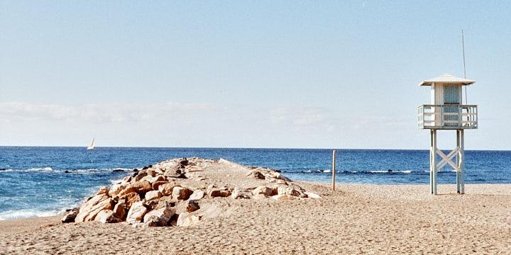 A colour film photo looking out to sea. In the foreground is a deserted beach with a rocky outcrop and a lifeguard lookout hut on the right. On the sea horizon, there is a sailing boat.