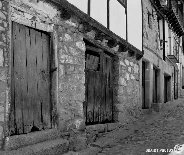A black-and-white film photo looking along a row of terraced houses on the left of a cobbled street. The first two timber doors are very old and have decayed along the bottom.