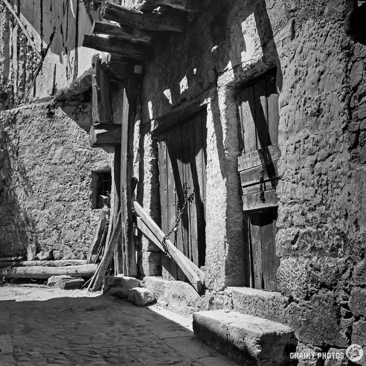 A black-and-white film photo of two very old timber doors, chained and secured. It looks like animals may have been kept here; the doors do not appear to be used regularly.