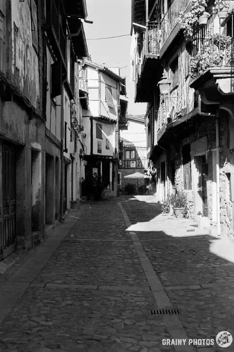 A black-and-white film photo of the main street through the old quarters of Mogarraz. Three-storey stone and timber-framed houses are on both sides of the cobbled street. The upper storeys of some of the houses cantilever out into the street.