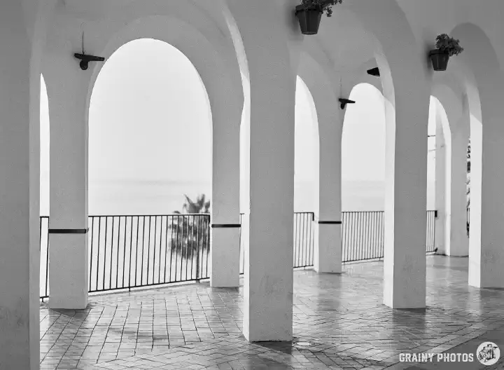A black and white film photo looking out to sea through the white arches on the Plaza Balcon De Europa. The photo was taken early, on a misty morning when no people were about.