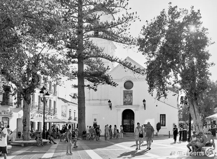 A black and white film photo of Iglesia de El Salvador in Nerja. A plaza is in front of the church with several trees. People walking around.