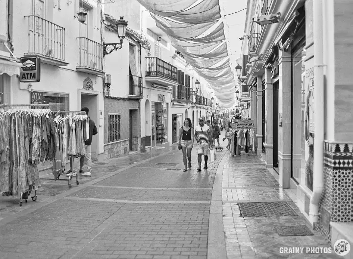 A black and white film photo of a pedestrianised block-paved shopping street in Nerja. A few tourists/shoppers are wandering around. Sun shades are stretched above the street to provide shade.