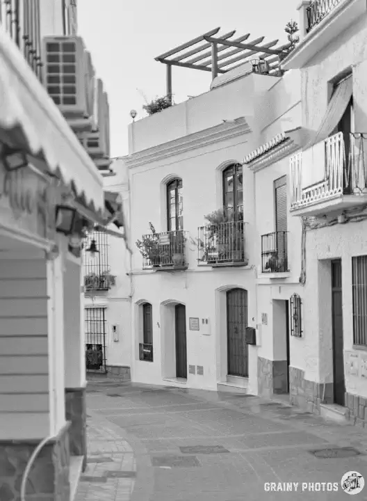 A black and white film photo of a narrow block-paved street in Nerja with white houses on both sides.