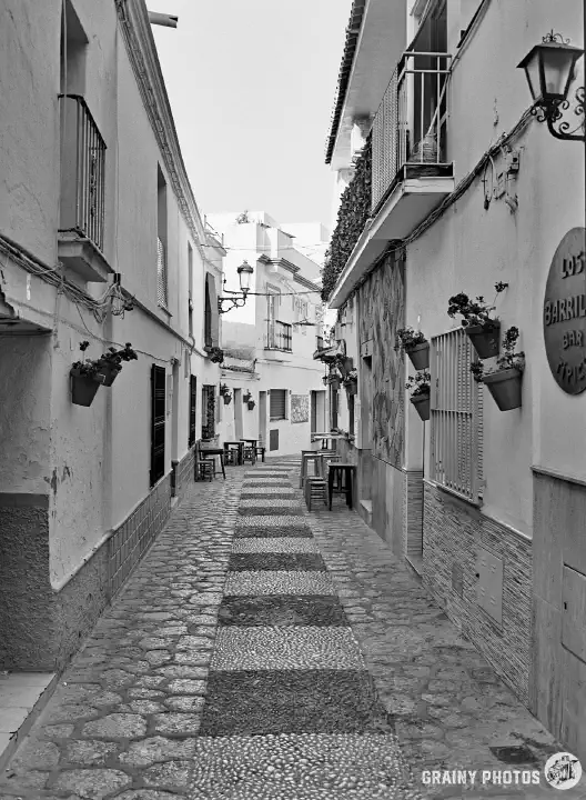 A black and white film photo of an empty, narrow side street in Nerja. Cafe tables are out in the street on both sides in the distance.