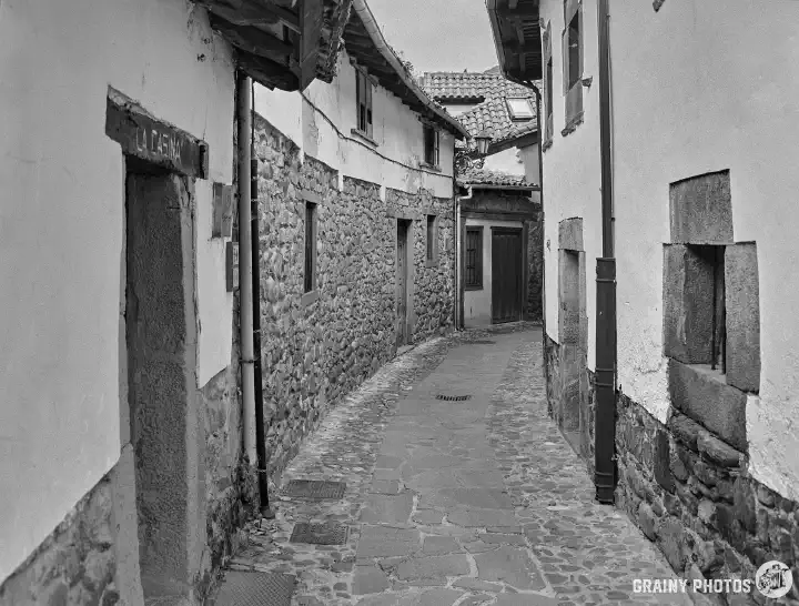 A black-and-white film photo of a cobbled alley curving to the right, with low stone houses on both sides.