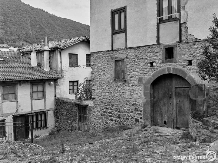 A black-and-white film photo of houses by a cobbled, somewhat overgrown street. The house on the right is larger with a big timber double entrance door.