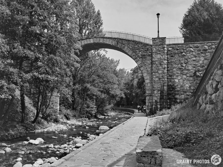 A black-and-white film photo of a footbridge over the Quiviesa River. A footpath follows the right bank of the river.