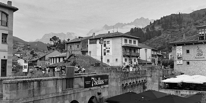 A black-and-white film photo in the centre of Potes showing the road bridge across the river. Beyond are townhouses with mountains in the mist in the background.