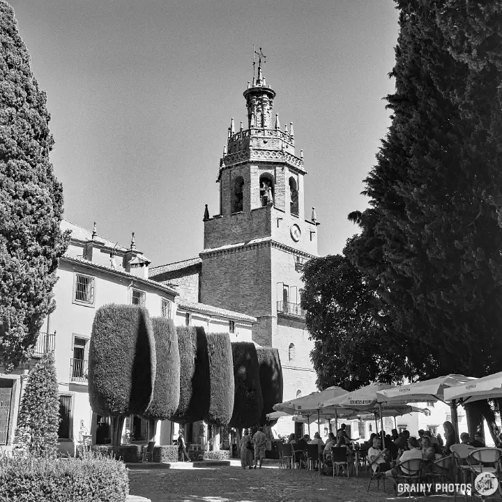 A black-and-white film photo of the Church of Santa María la Mayor. The photo is taken from the square in front of the church. On the right-hand side, in the shade of trees, is a street cafe with tables and chairs and parasols.