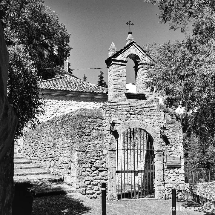 A black-and-white film photo of the restored ruins of the La Ermita de San Miguel. Just the stone walls with the main entrance and a small belfry are standing.