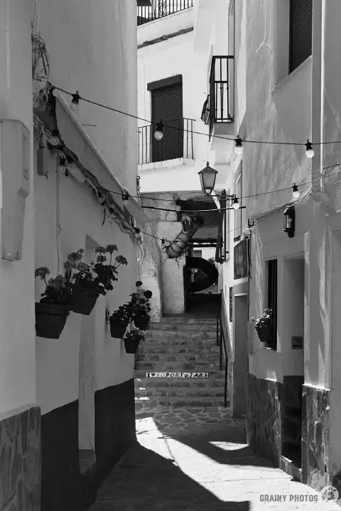 A  black-and-white film photo of narrow streets amongst white houses. Stone steps lead up to a tinao... with a ... what is that in the tinao? All will be revealed in the next photo...